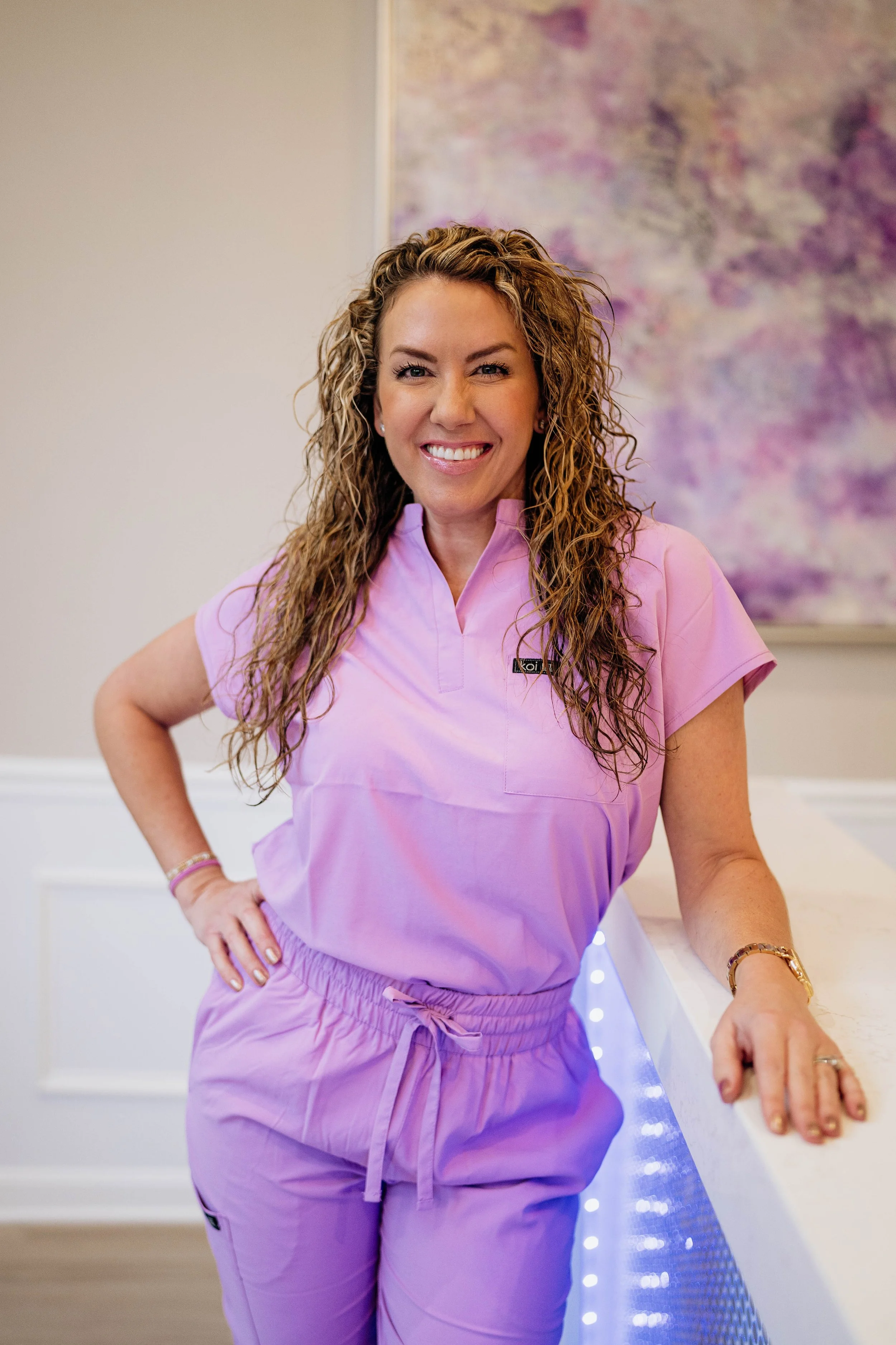 A woman with curly blonde hair smiling while wearing pink medical scrubs, standing indoors near a wall with abstract purple artwork and LED strip lights.