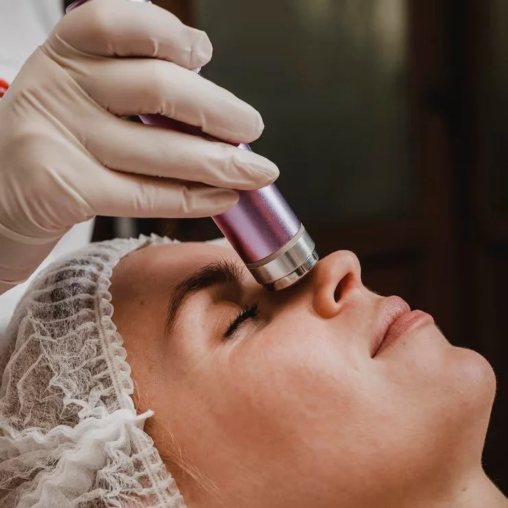 Skincare professional performs a facial treatment with a violet device on a woman's face, wearing a hairnet and gloves.