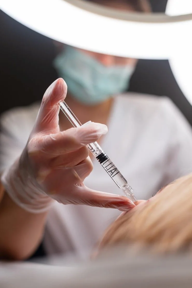 A medical professional wearing gloves and a face mask administering an injection into a patient's arm with a syringe.