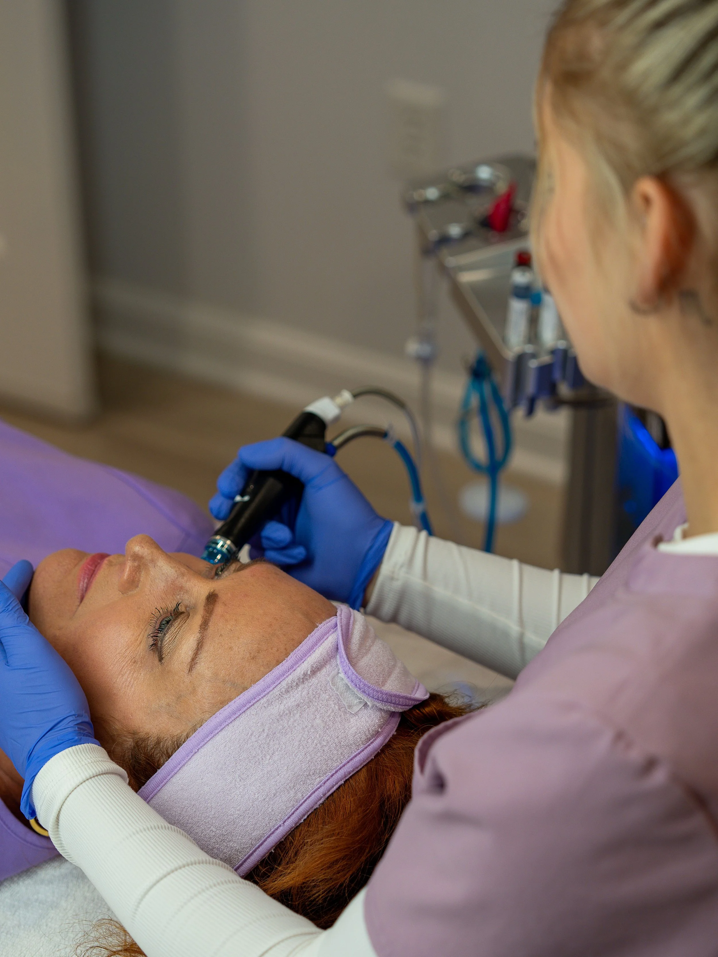 A woman lying on a medical bed with her eyes closed, receiving a cosmetic or skincare treatment from a healthcare professional wearing purple scrubs and blue gloves. The professional is using a handheld device on the woman's face near her lips.