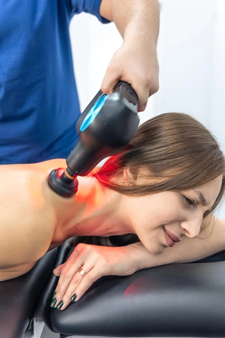 A woman lying face down on a treatment table, receiving a laser therapy treatment on her neck from a medical professional.