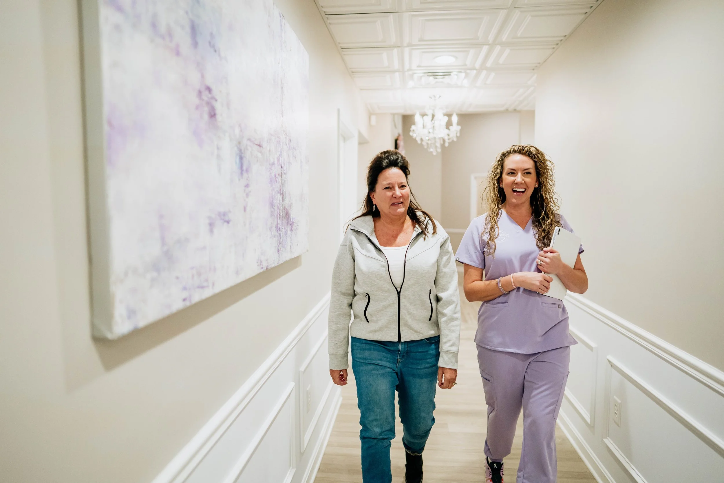 Two women walking down a hallway; one is in medical scrubs, smiling, holding a white tablet, the other is in casual clothes, listening with earphones.