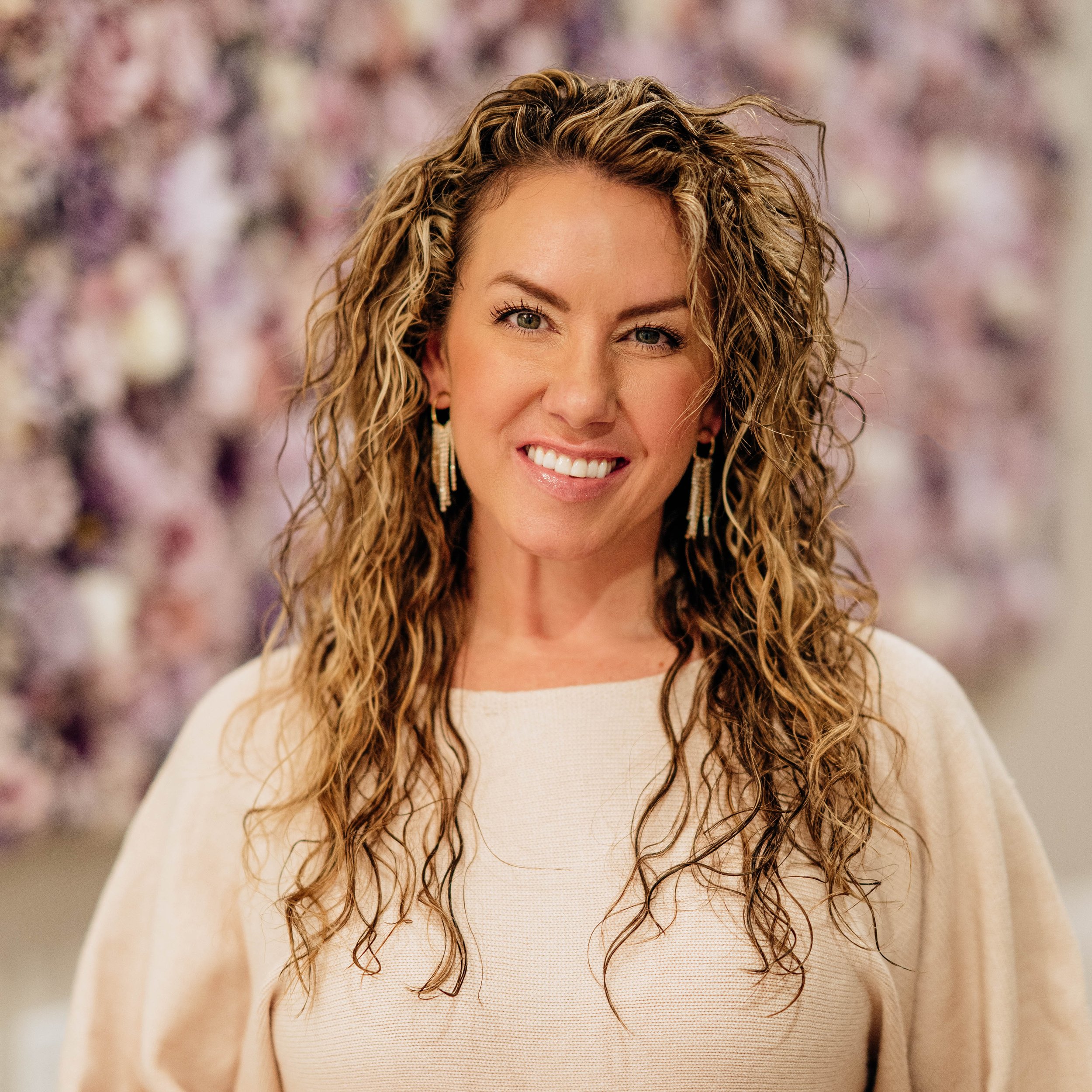 Close-up of a woman with curly blond hair, smiling, wearing earrings and a cream-colored top.