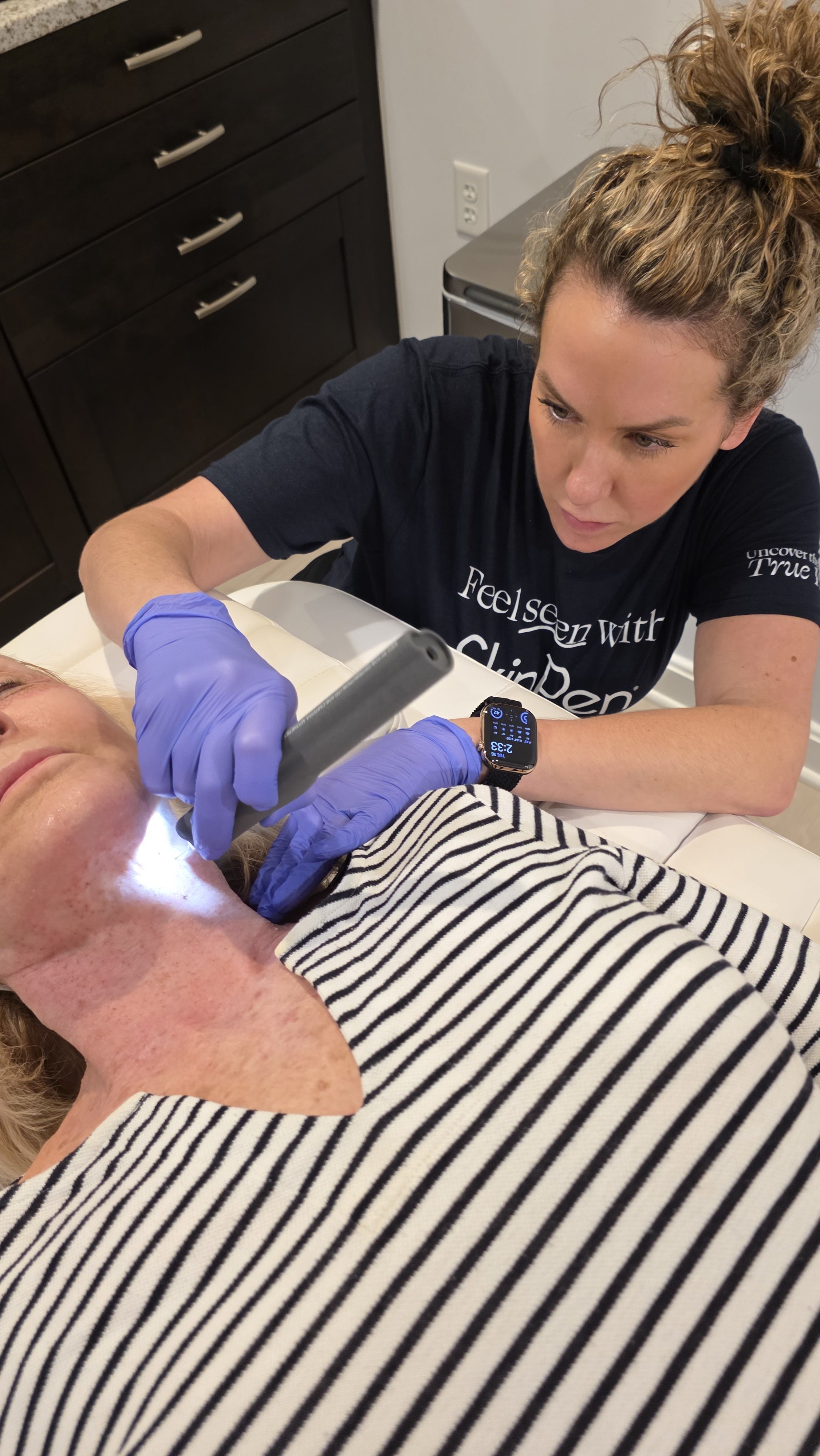 A healthcare professional with curly hair wearing a black T-shirt and blue gloves is examining a patient with a medical device near her mouth. The patient is lying on a bed wearing a striped shirt.