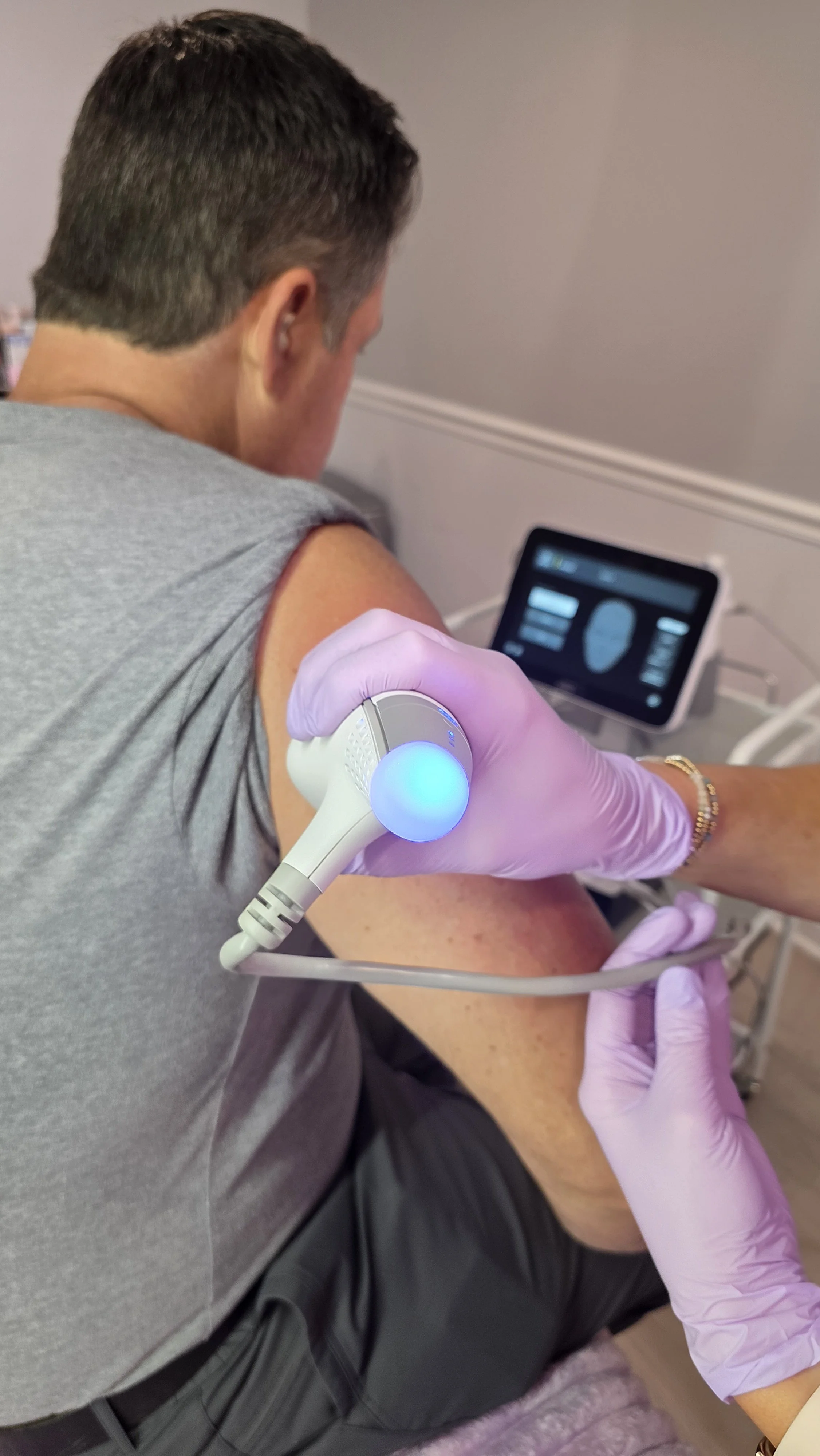 A person receiving a medical ultrasound scan on their upper arm while seated, with a healthcare professional wearing purple gloves operating the ultrasound device. A monitor in the background displays the ultrasound imagery.