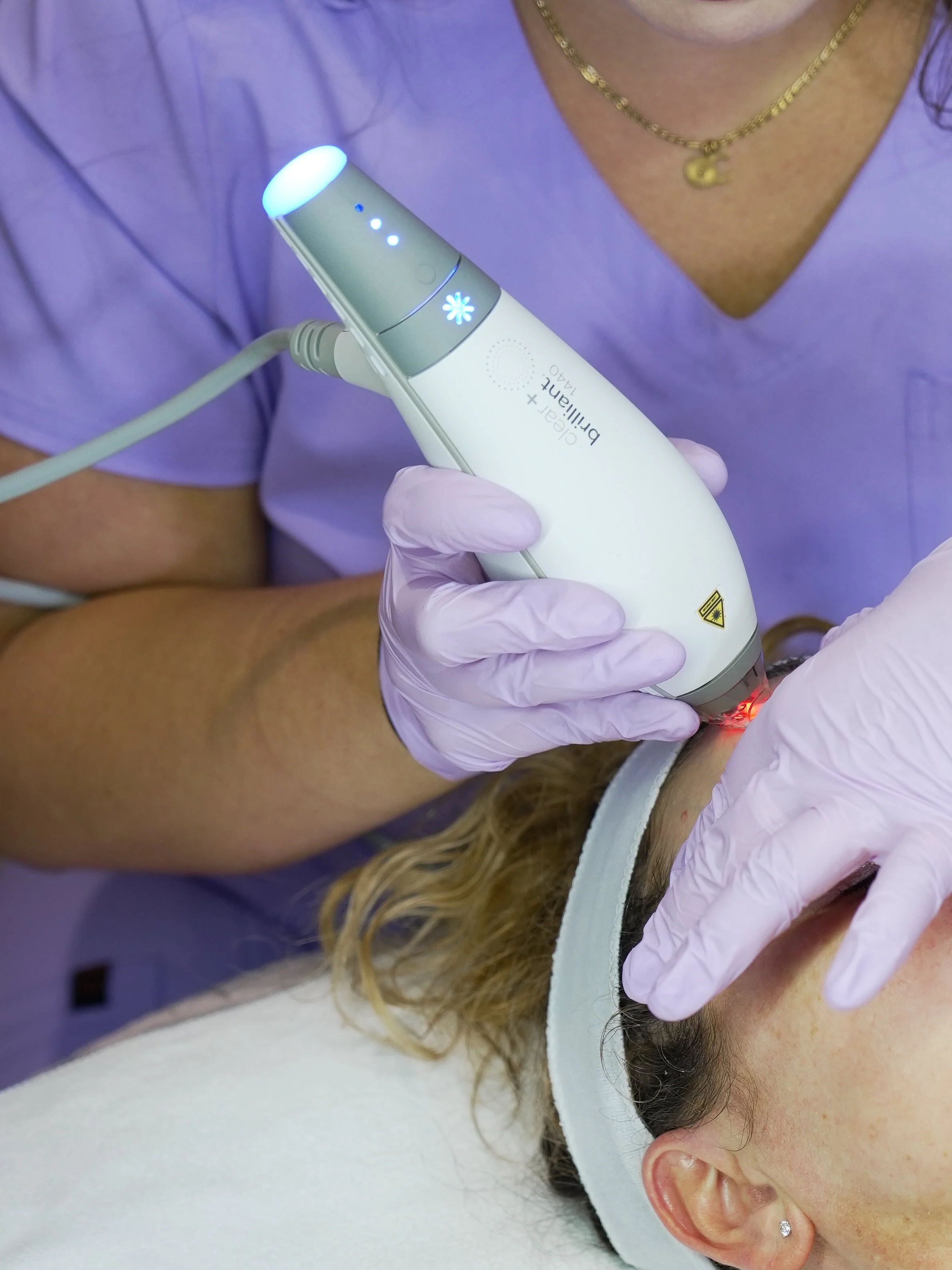 A person receiving a cosmetic laser treatment on their forehead, wearing a white headband and pink gloves, with a healthcare professional holding the laser device.