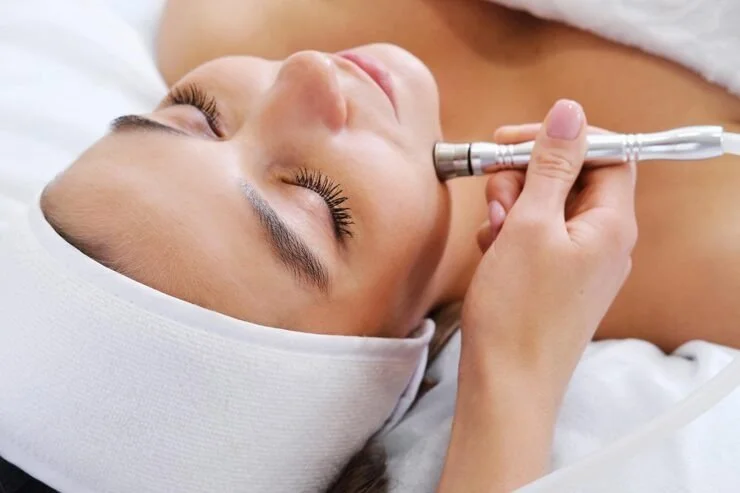 A woman lying down with her eyes closed, receiving a facial treatment with a skincare device held by a professional