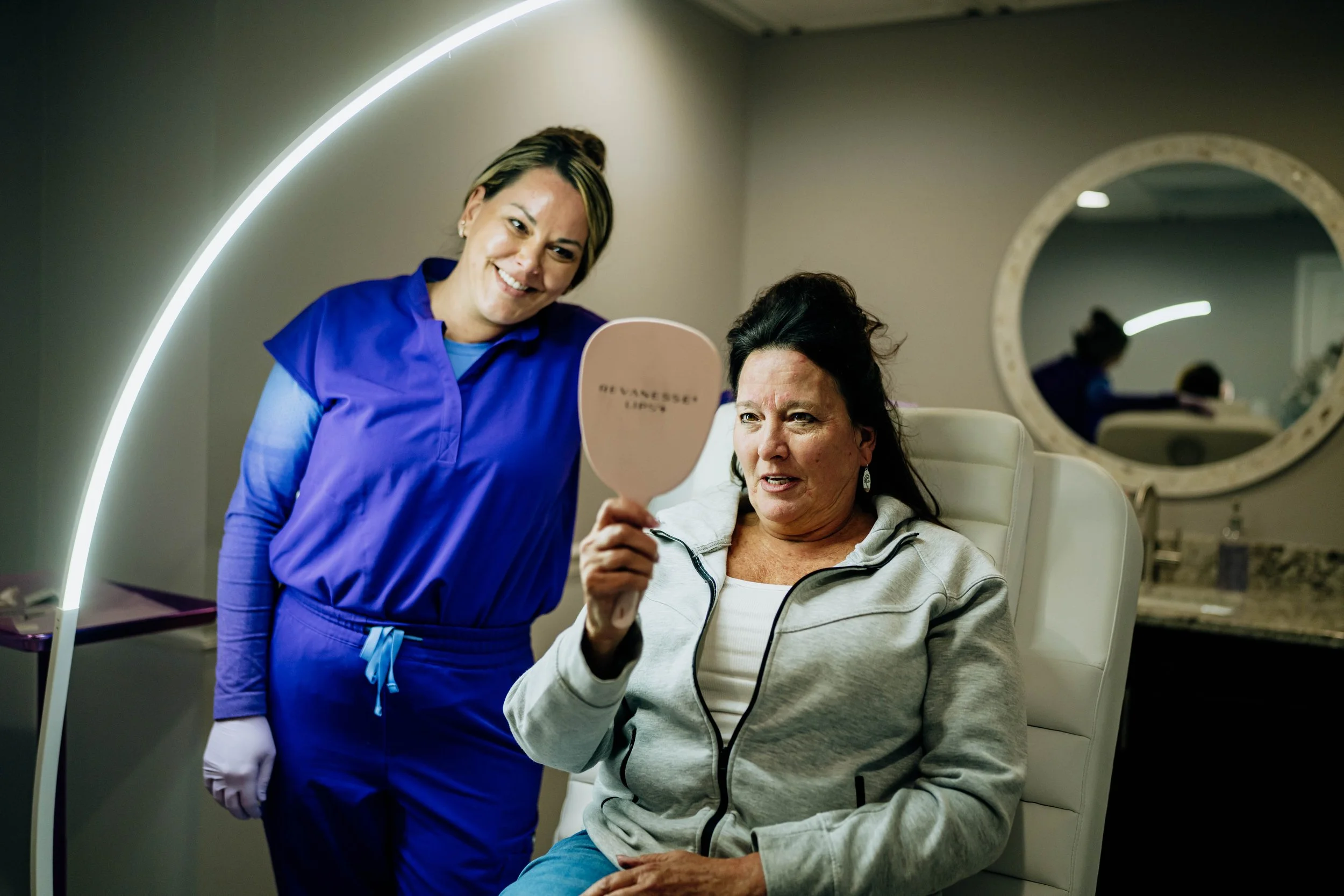 A healthcare worker in blue scrubs smiling while holding a mirror and helping a woman sit in a medical chair, with a mirror on the wall behind them reflecting their image.