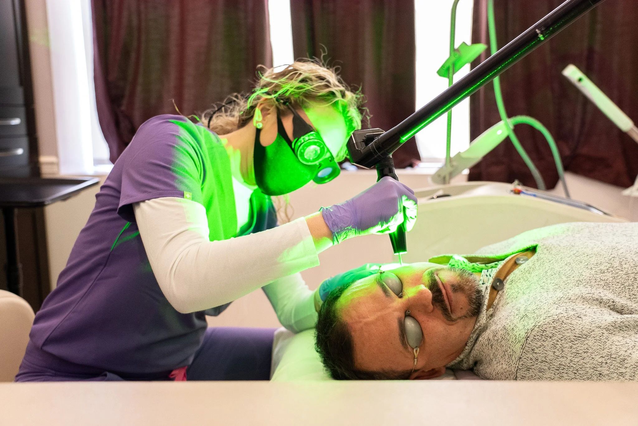 A woman with curly hair, wearing a face mask and purple scrubs, is using a green laser or light device on a man's forehead as he lies on a medical bed. The setting appears to be a medical or clinic environment.