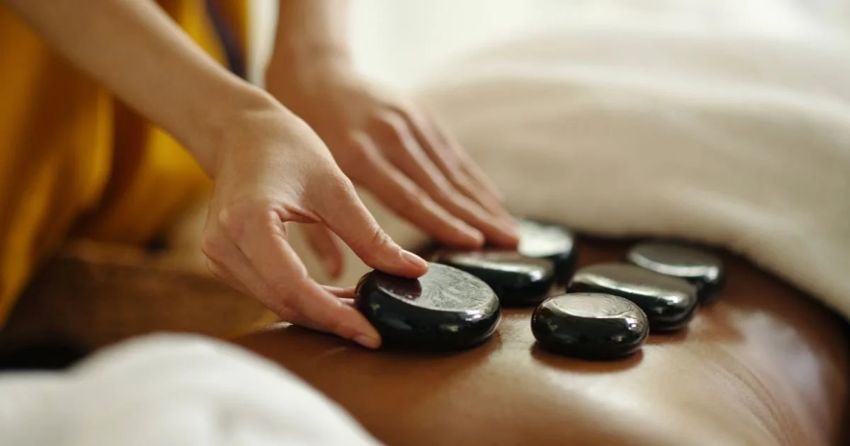 A person placing smooth, black massage stones on their back during a spa treatment.