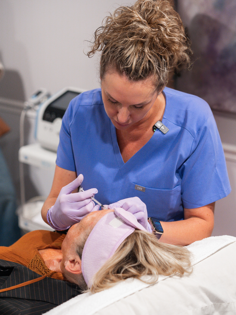 A nurse in blue scrubs preparing to give an injection to an elderly woman lying in a medical bed.