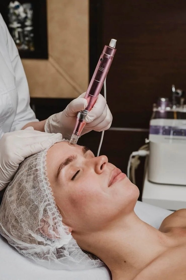 A woman lying down with her eyes closed, receiving a facial treatment with a cosmetic device held by a professional wearing gloves.