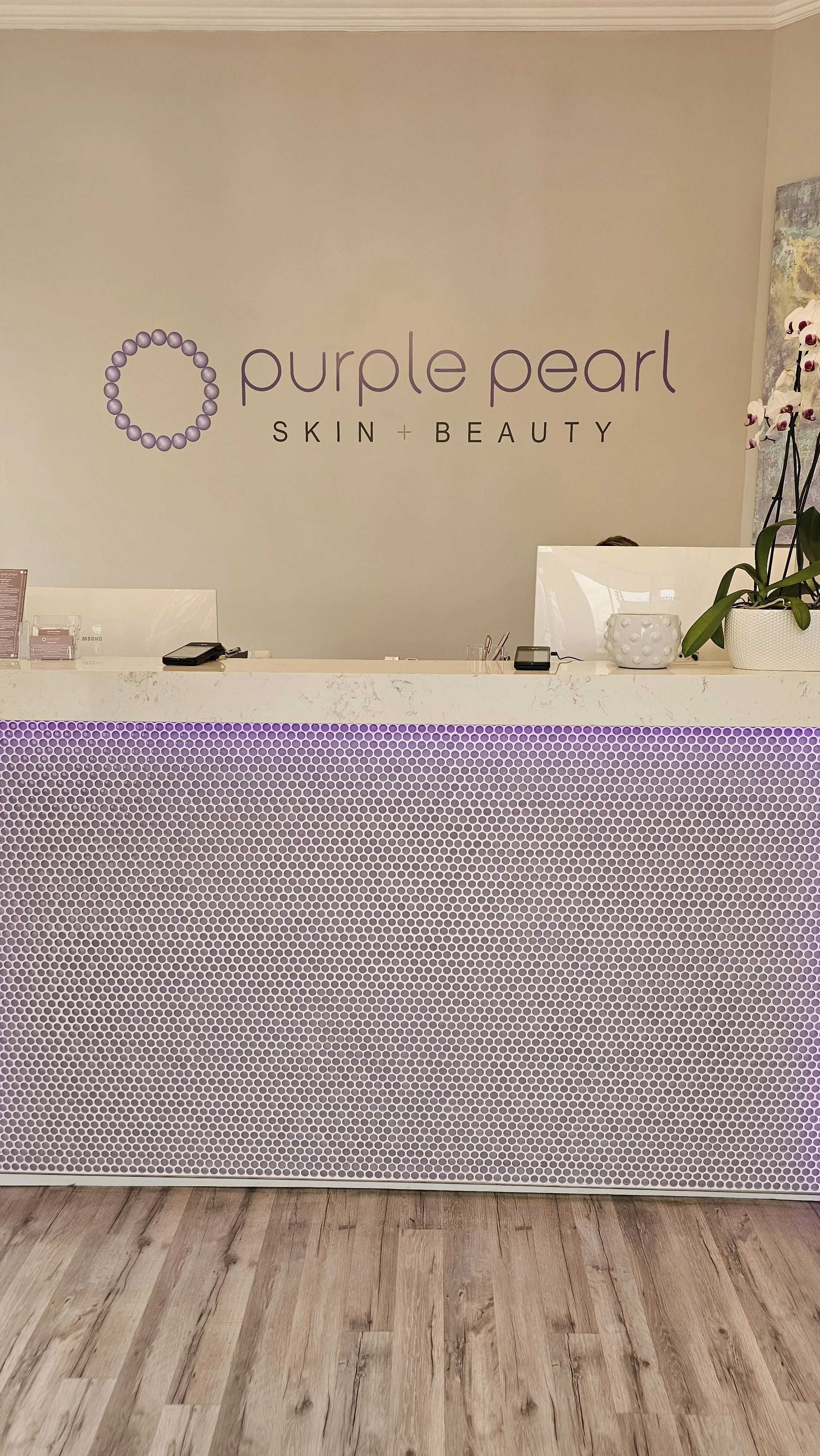 Reception area of a skin and beauty clinic with a purple pearl-themed decor, a reception desk with a purple honeycomb pattern, a potted orchid plant, and a beige wall with the clinic's name and logo.