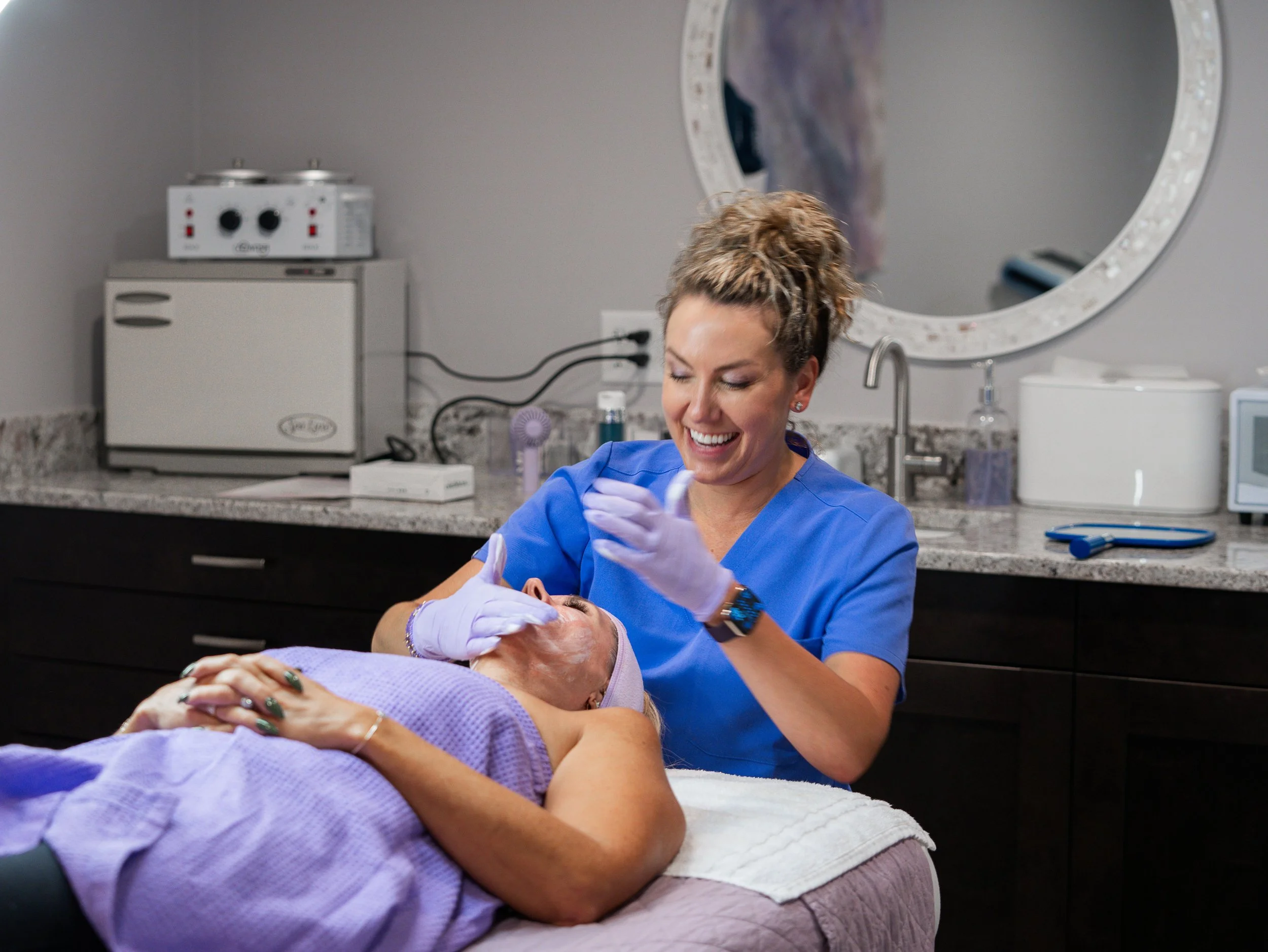 A healthcare worker in blue scrubs and gloves smiling as she helps an elderly patient lying down on a medical examination bed in a clinic or hospital setting.