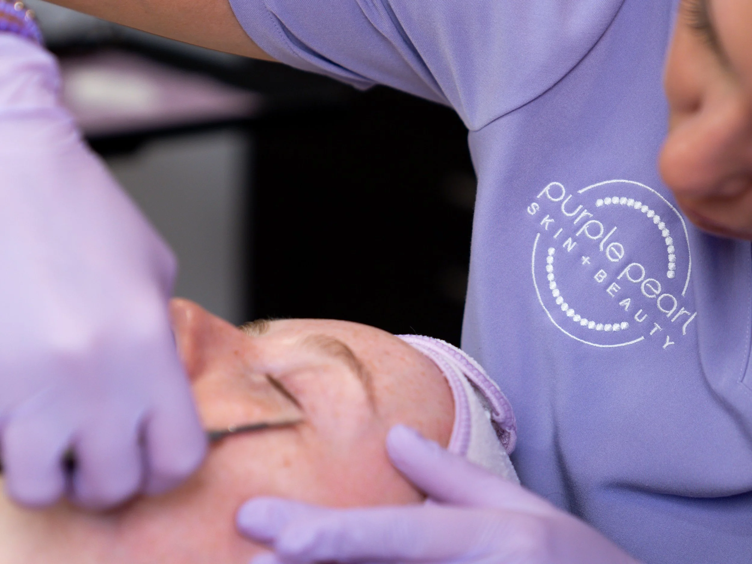 Close-up of a healthcare professional's hand in purple glove, administering an injection to an elderly person's arm with visible wrinkles, wearing a pink sleeve. The healthcare worker's shirt is light purple with a logo that reads 'Pureology Skin + Beauty'.