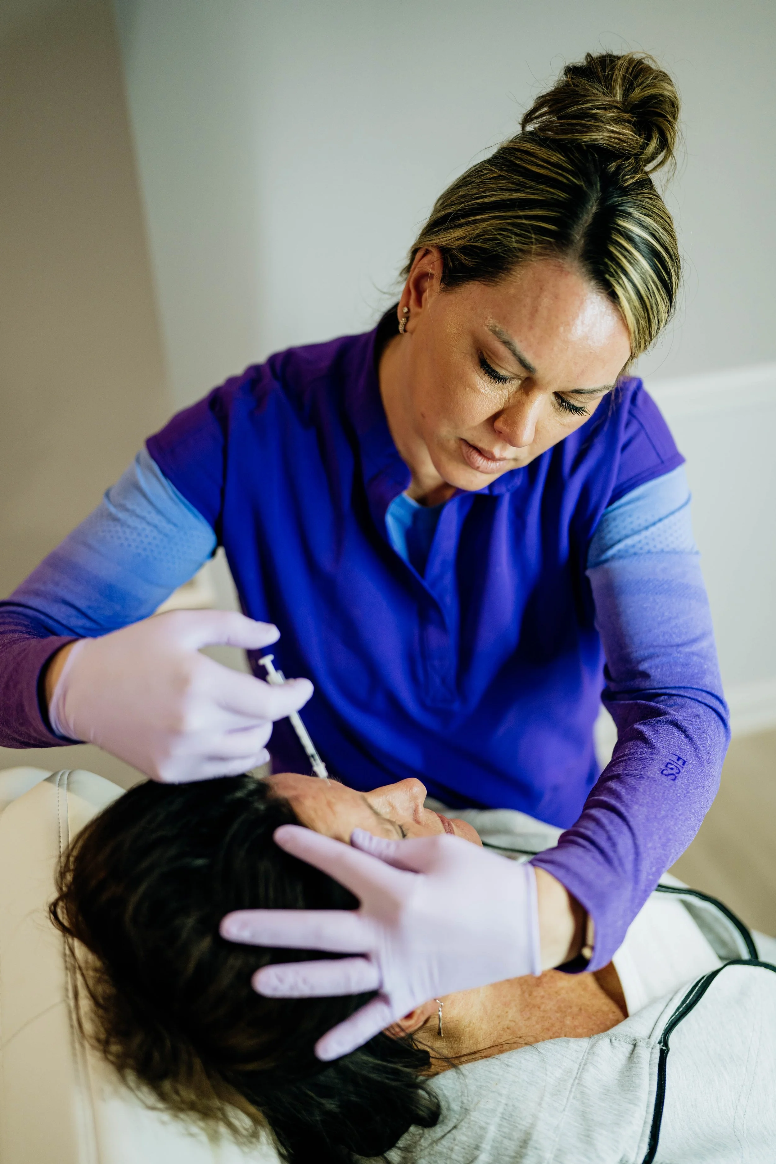 Medical professional administering a facial injection to a patient lying on a bed, with another person supporting the patient supportively.