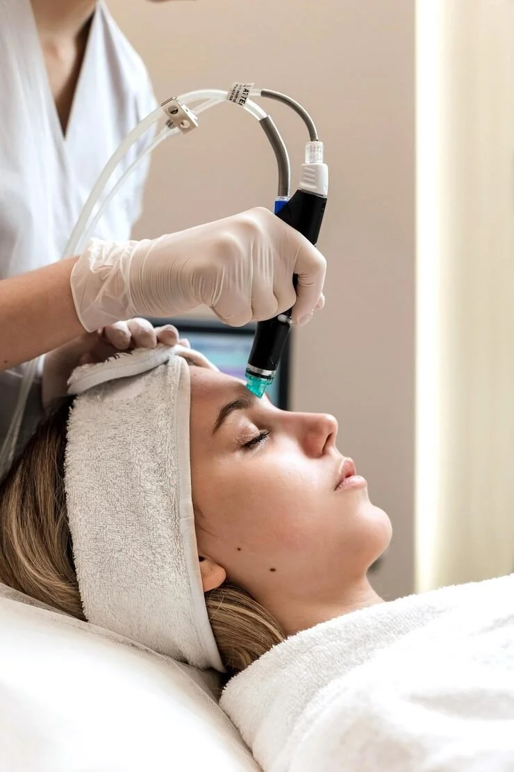 A woman receiving a facial treatment with a handheld device at a spa or clinic, lying down with her eyes closed and head supported by a towel.