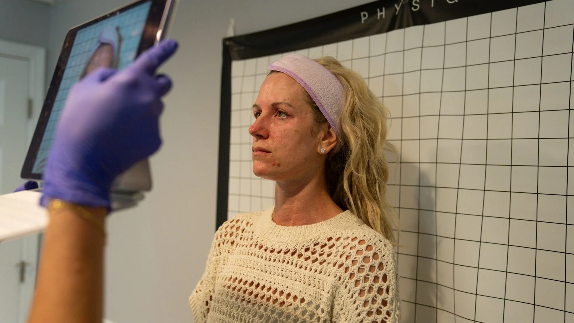 A woman with blonde, curly hair and a pink headband stands against a grid background while a person in purple gloves takes a photo of her with a tablet.