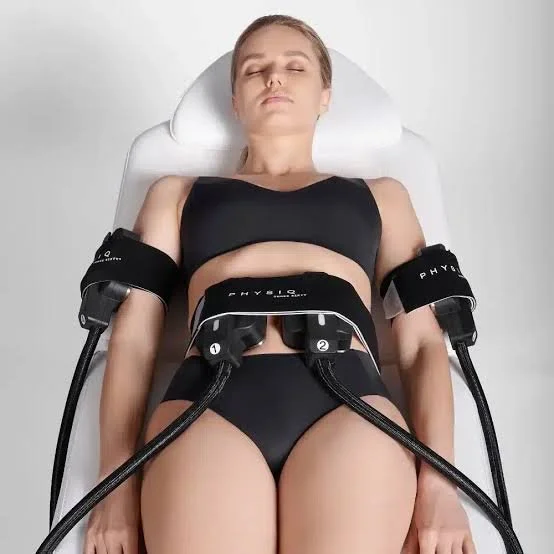 A woman lying on a treatment table wearing black workout attire, connected to two pieces of medical equipment on her arms and abdomen.