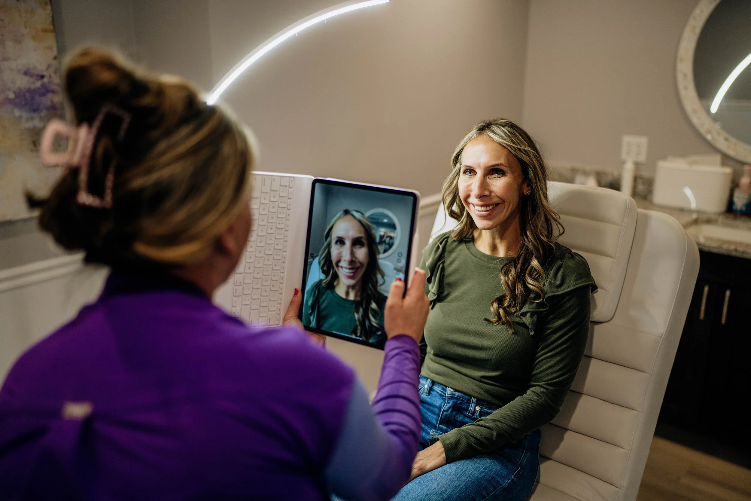 A woman with long, wavy hair and a green top smiling while sitting in a chair and having her photo taken by another person holding a tablet. The person taking the photo has blonde hair tied back with pink hair clips and is wearing a purple top. The background features a mirror, a wall with a modern light strip, and some household items.