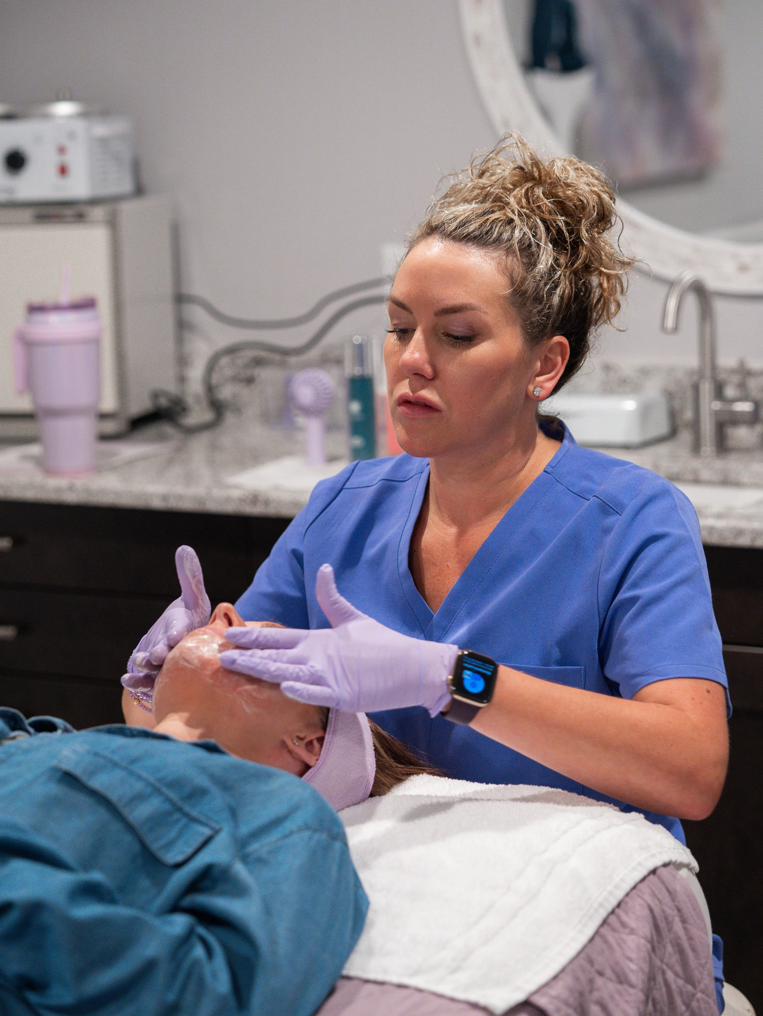 A healthcare professional, wearing a blue uniform and purple gloves, attentively cares for a patient lying on a medical bed. The healthcare worker is focused, possibly preparing to perform a medical procedure or examination. The background includes medical equipment, bottles, and a mirror.