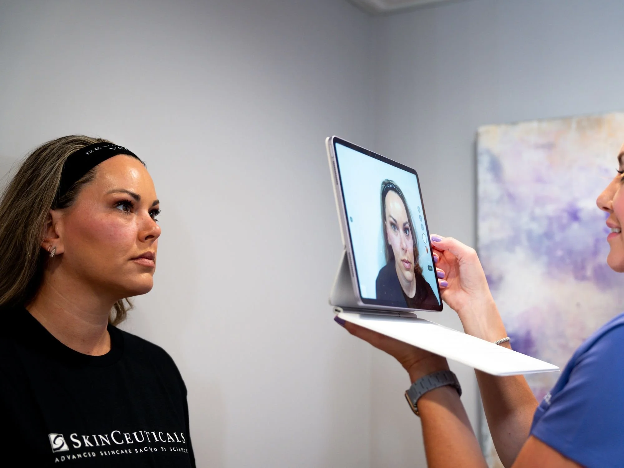 A woman with a black headband and earrings looks at a portrait on a tablet held by another person. The woman wears a black shirt with the logo 'SkinCeuticals'. The person holding the tablet is wearing a blue shirt and a watch.