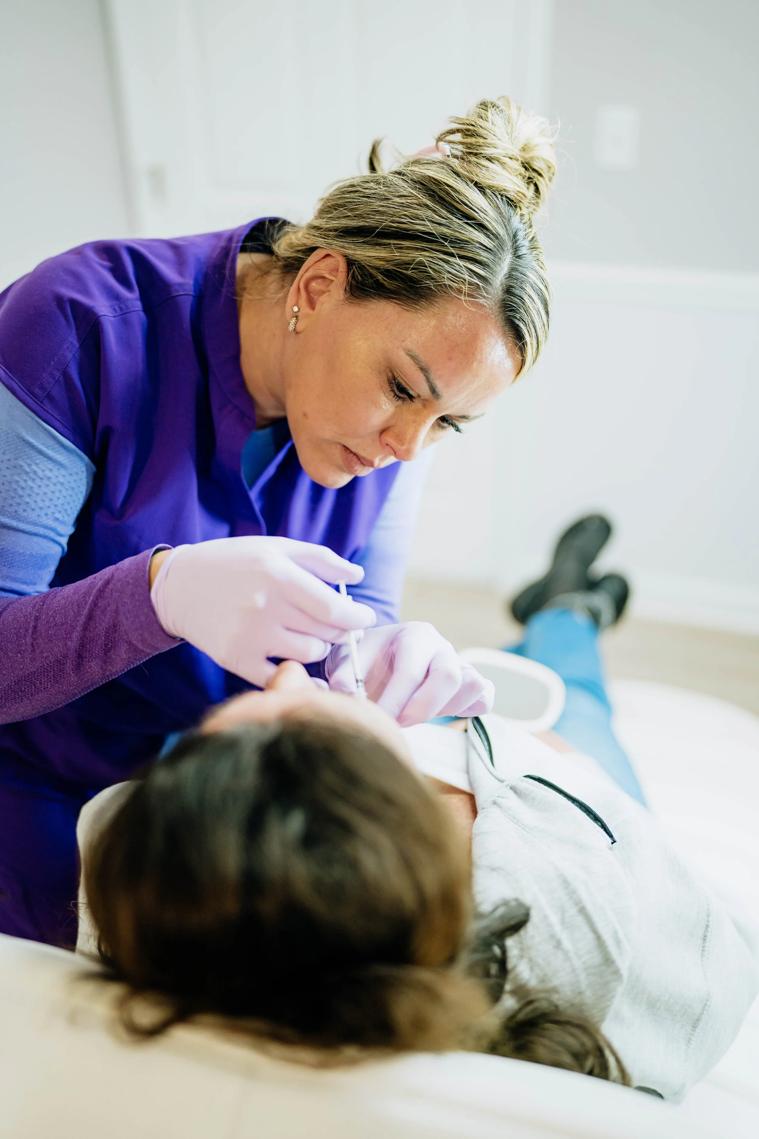 A healthcare professional, possibly a nurse, wearing purple scrubs and gloves, is tending to a patient lying on an examination table. The scene appears to be in a medical setting, such as a clinic or hospital.
