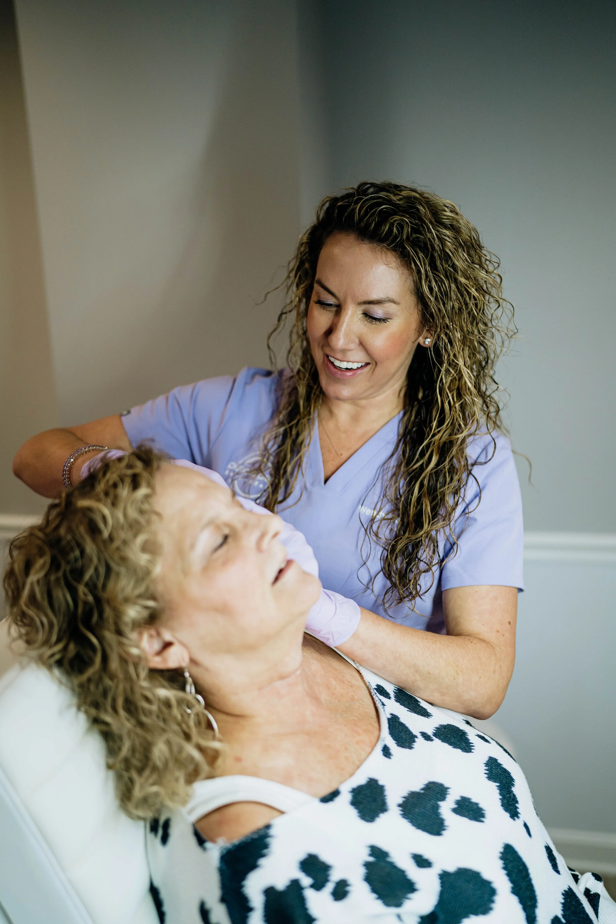 A woman with curly blonde hair wearing a white and black leopard print top is lying back with her eyes closed, while a healthcare professional with long, curly hair in scrubs looks down and smiles at her.