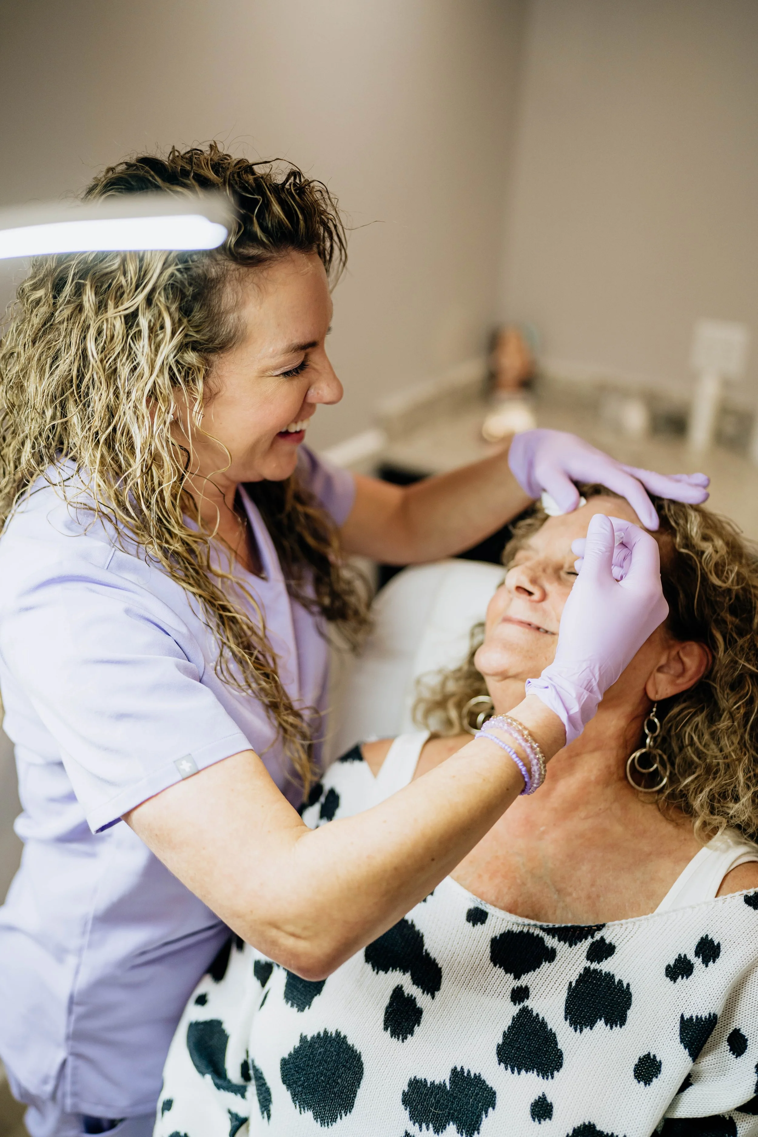 A healthcare worker with curly blonde hair, wearing a purple uniform and gloves, smiling while performing a procedure on an older woman with curly blonde hair, who is seated and wearing a black and white animal print top.