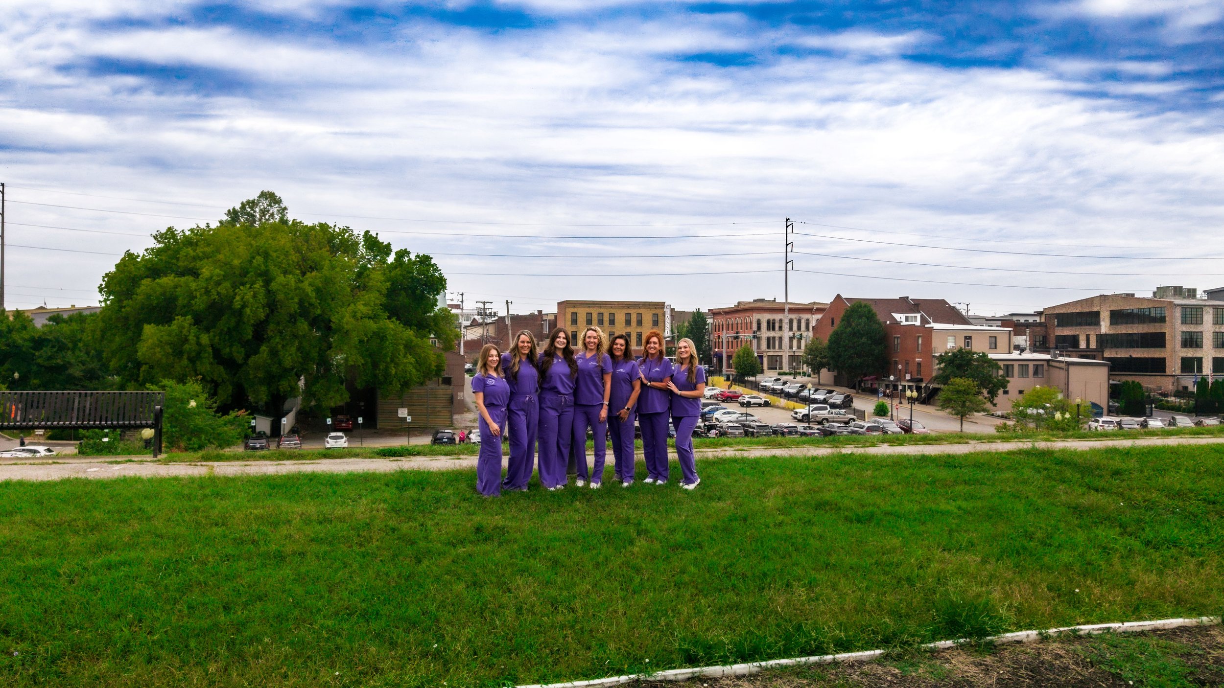 A group of seven women dressed in purple scrubs standing on a grassy area outdoors, with a cityscape and parking lot in the background under a cloudy sky.