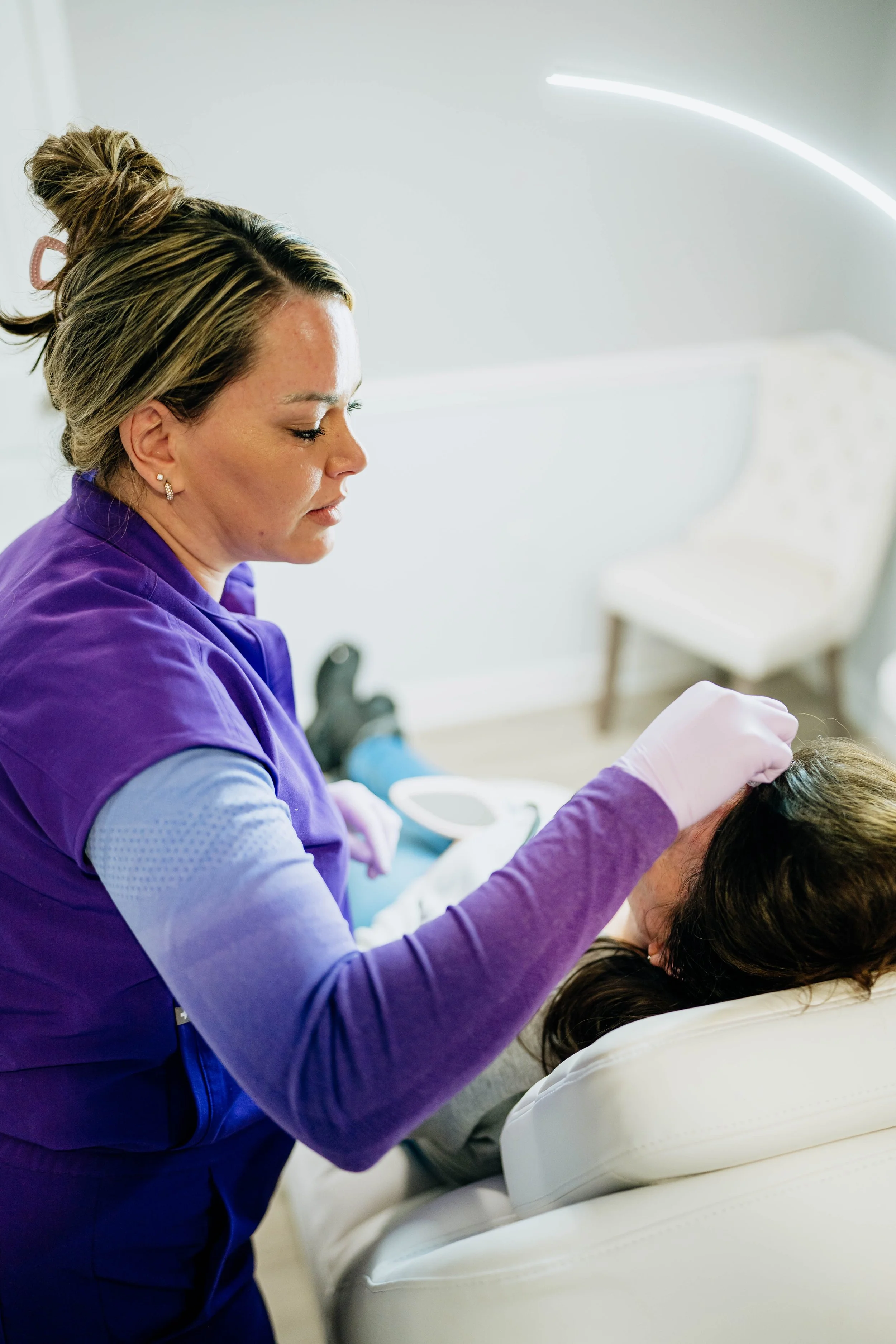 A nurse in purple scrubs and gloves is tending to a patient who is lying in a white medical bed. The nurse appears focused, with her eyes closed, and is working on the patient's head or hair.