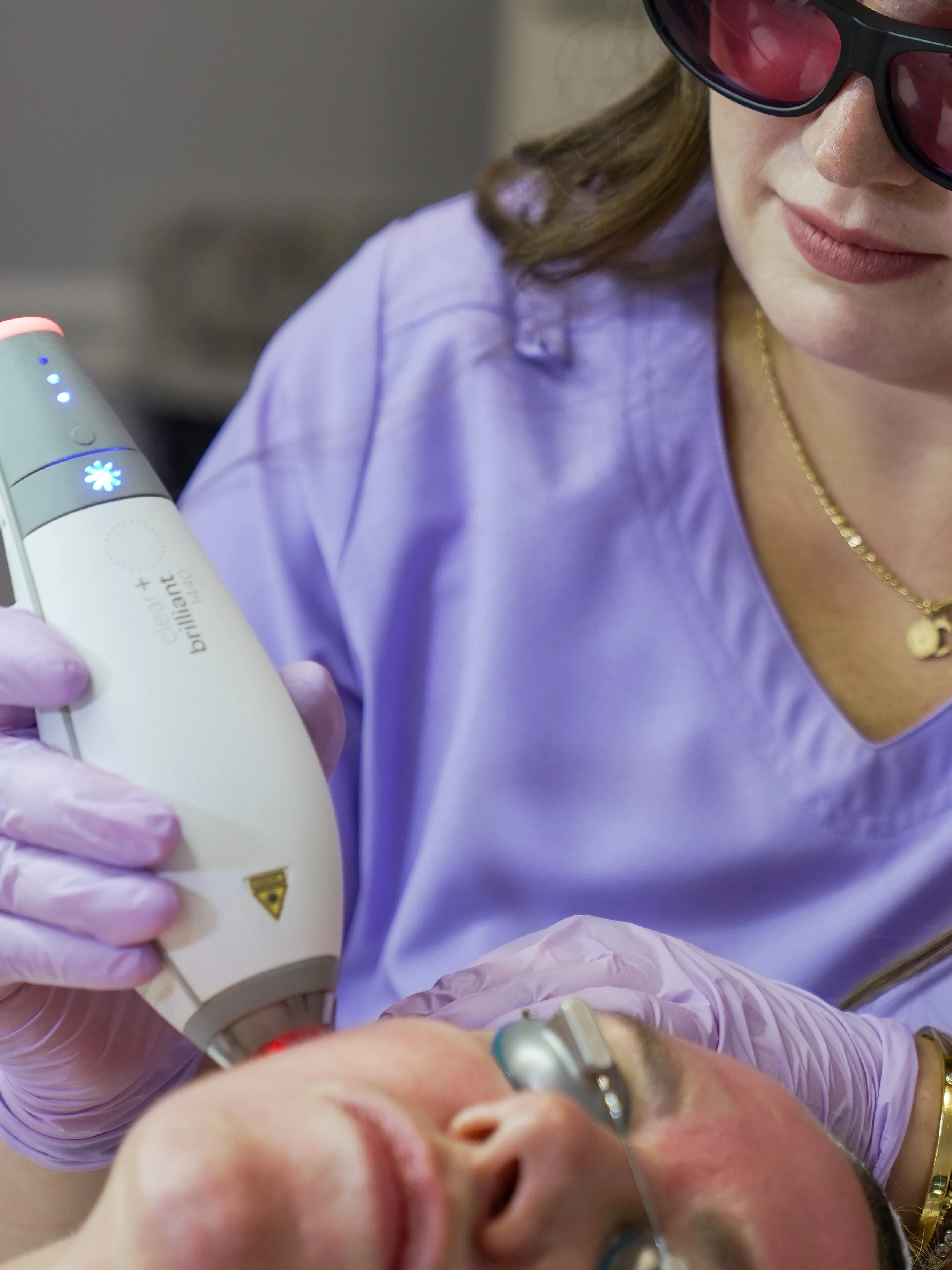 A healthcare worker wearing pink gloves and purple scrubs is holding an ultrasound device against a patient's forehead. The patient, with their eyes closed, has a nasal cannula providing oxygen, and has light-colored skin and hair.