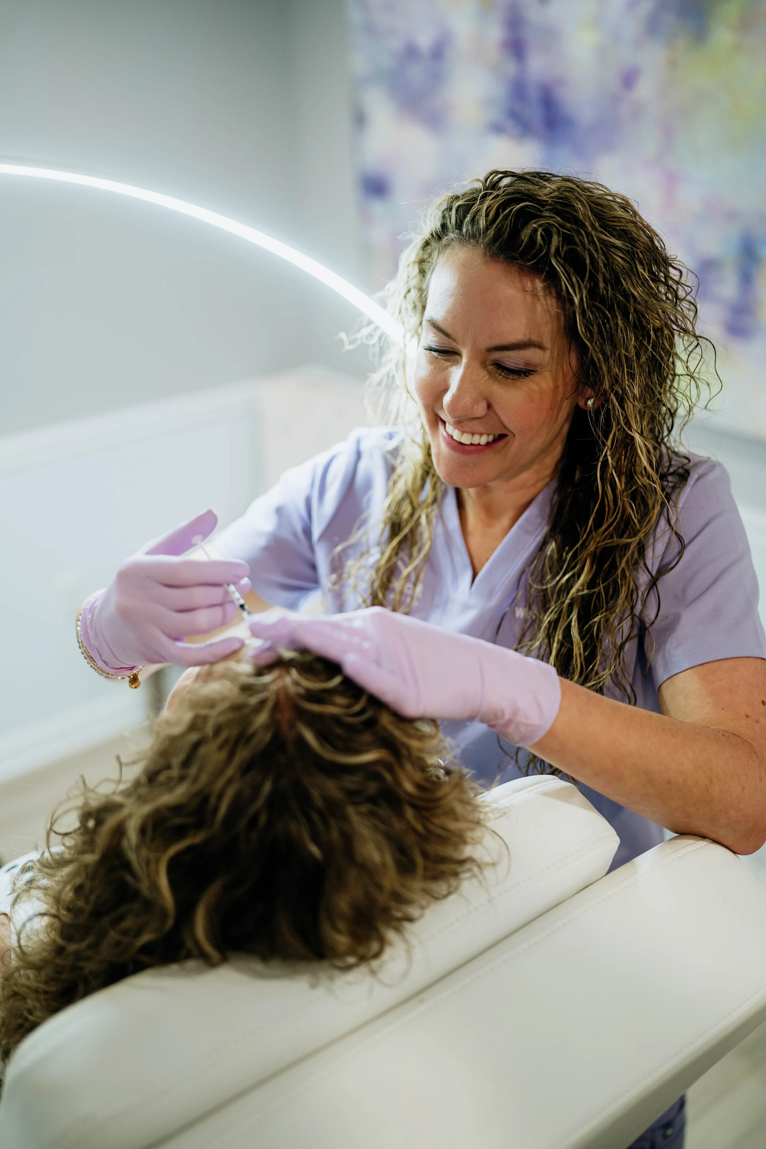 A woman with curly hair smiling while giving an injection to a patient lying on a medical examination table.