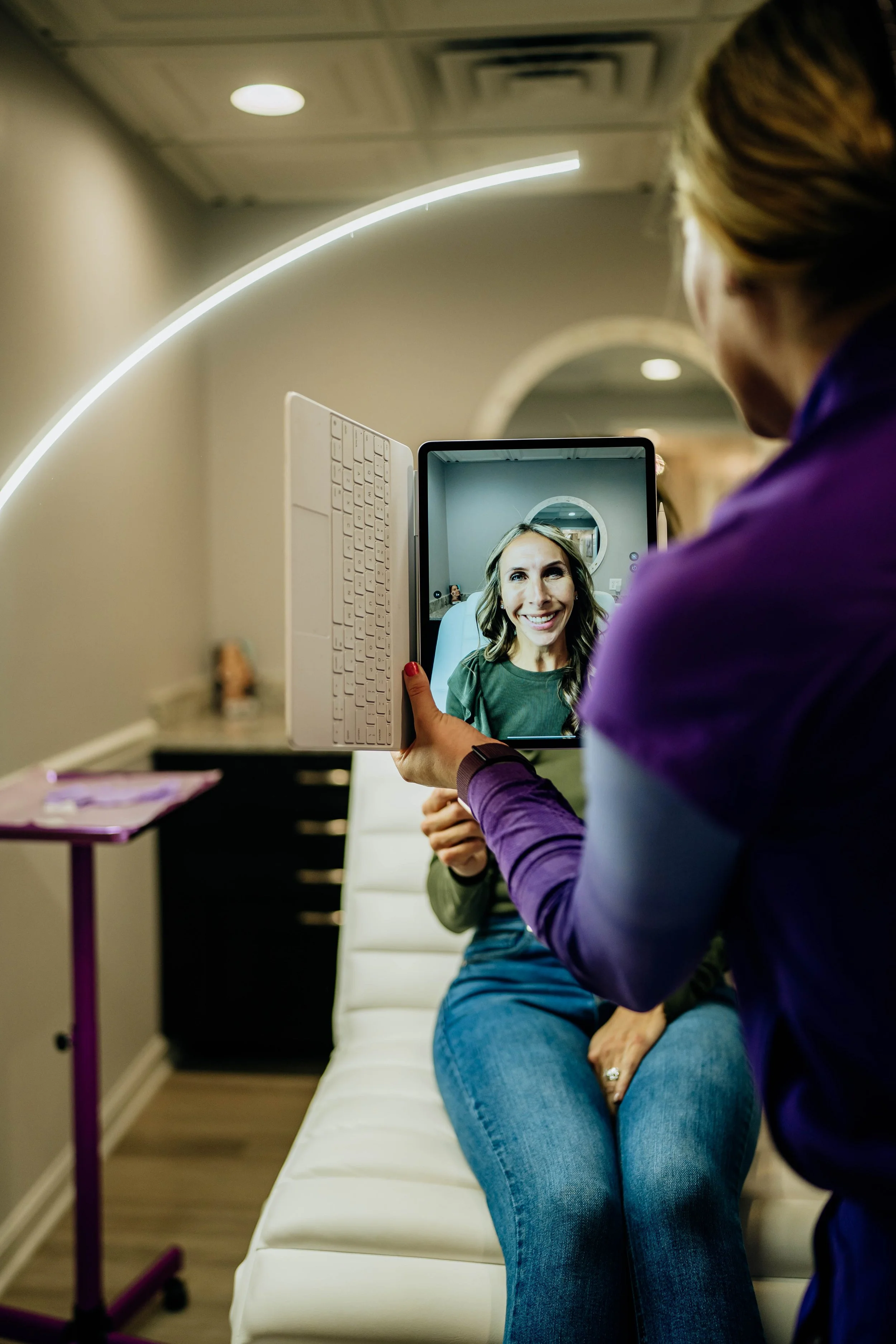 A woman sitting on a medical examination table smiling as she looks into a tablet held by a healthcare professional. The professional is using the tablet to view her reflection on the screen.