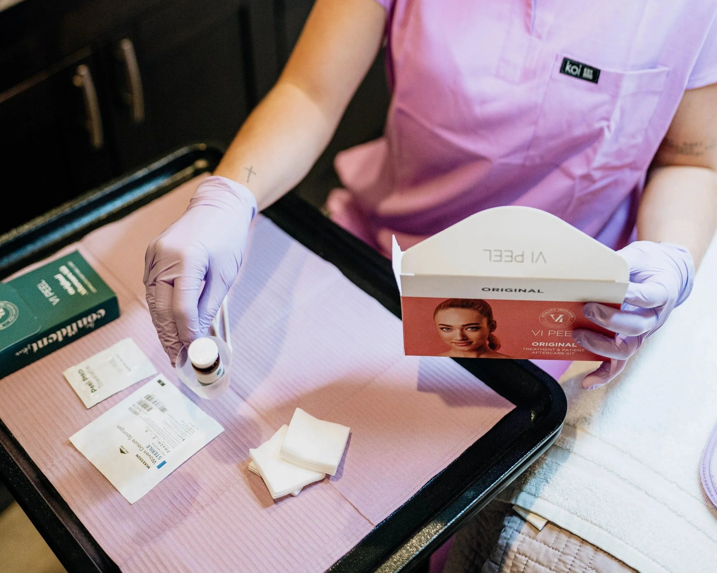Person wearing gloves handling medical supplies, including a box with a woman's face on it, medication bottles, and medical papers, on a pink table.