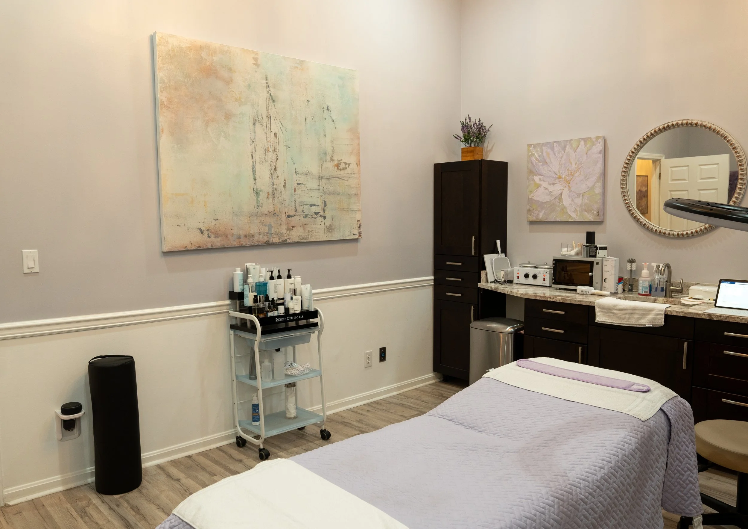 A room set up for medical or cosmetic treatments with two beds covered in purple quilts and folded white towels. There is a round mirror on the wall, a black cabinet with a lavender plant on top, and a countertop with medical equipment. A cart with skincare products and a black trash can are also visible.