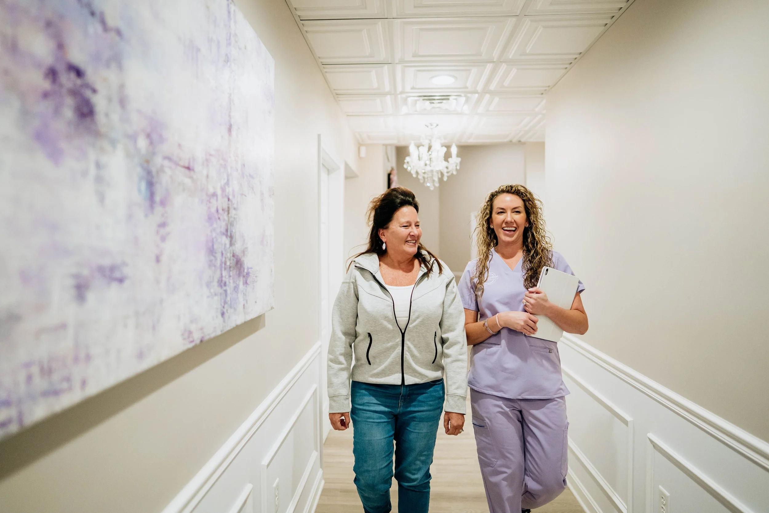 Two women, one in casual clothes and one in medical scrubs, walking down a hallway and smiling.