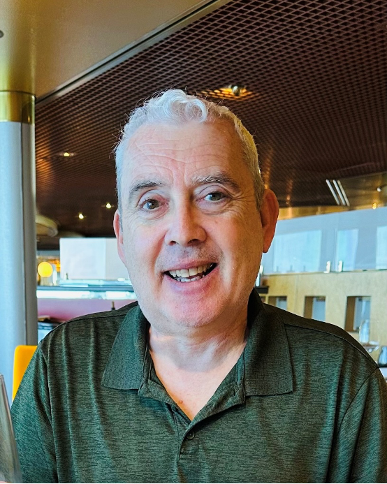 Ronald Keith smiling, wearing a dark green polo shirt, seated inside a restaurant or cafe with a modern interior.
