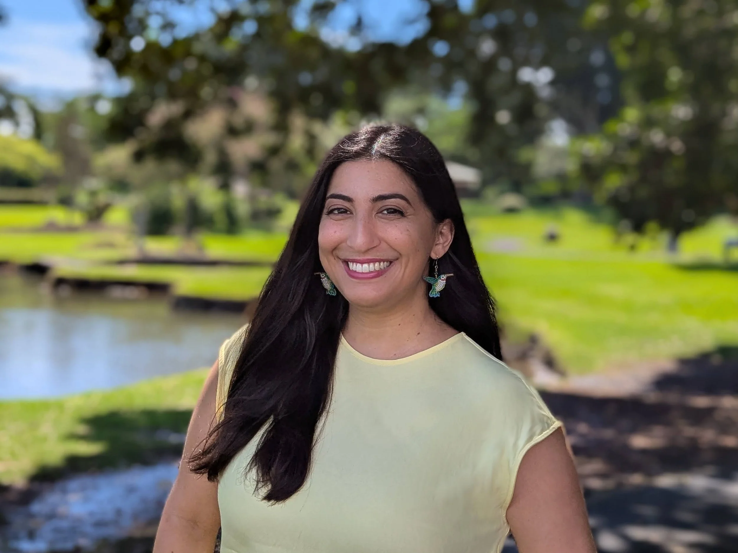 Samira Fatemi smiling, at Liliʻuokalani Gardens next to a body of water with greenery and trees in the background.