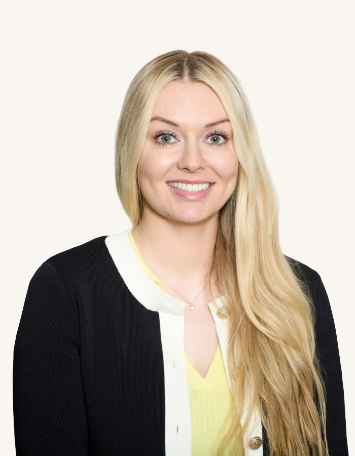 Portrait of a young woman with long blonde hair, smiling, wearing a black jacket over a light yellow top, against a plain white background.