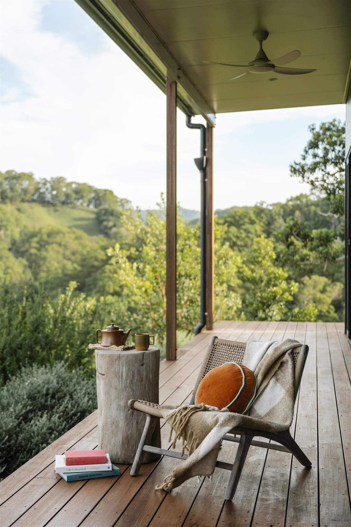 A porch with wooden flooring and a ceiling fan, overlooking a green landscape with trees and rolling hills. There is a chair with a blanket, a round orange cushion, a stump used as a table with a teapot and mug, and a few books on the floor.