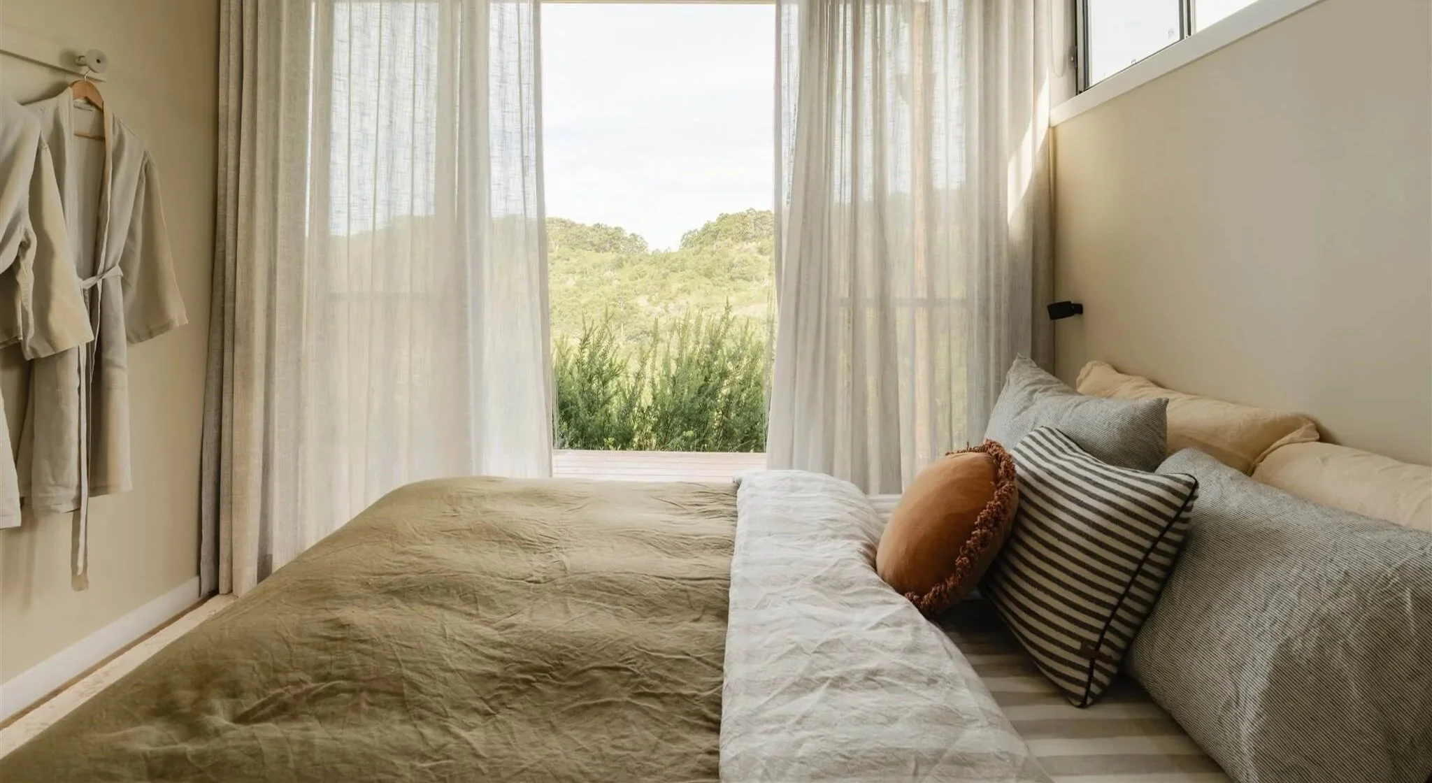 The single bedroom at Marys at Sunshine Coast Hinterland with large window curtains, a bed with multiple pillows, and a view of green hills outside.