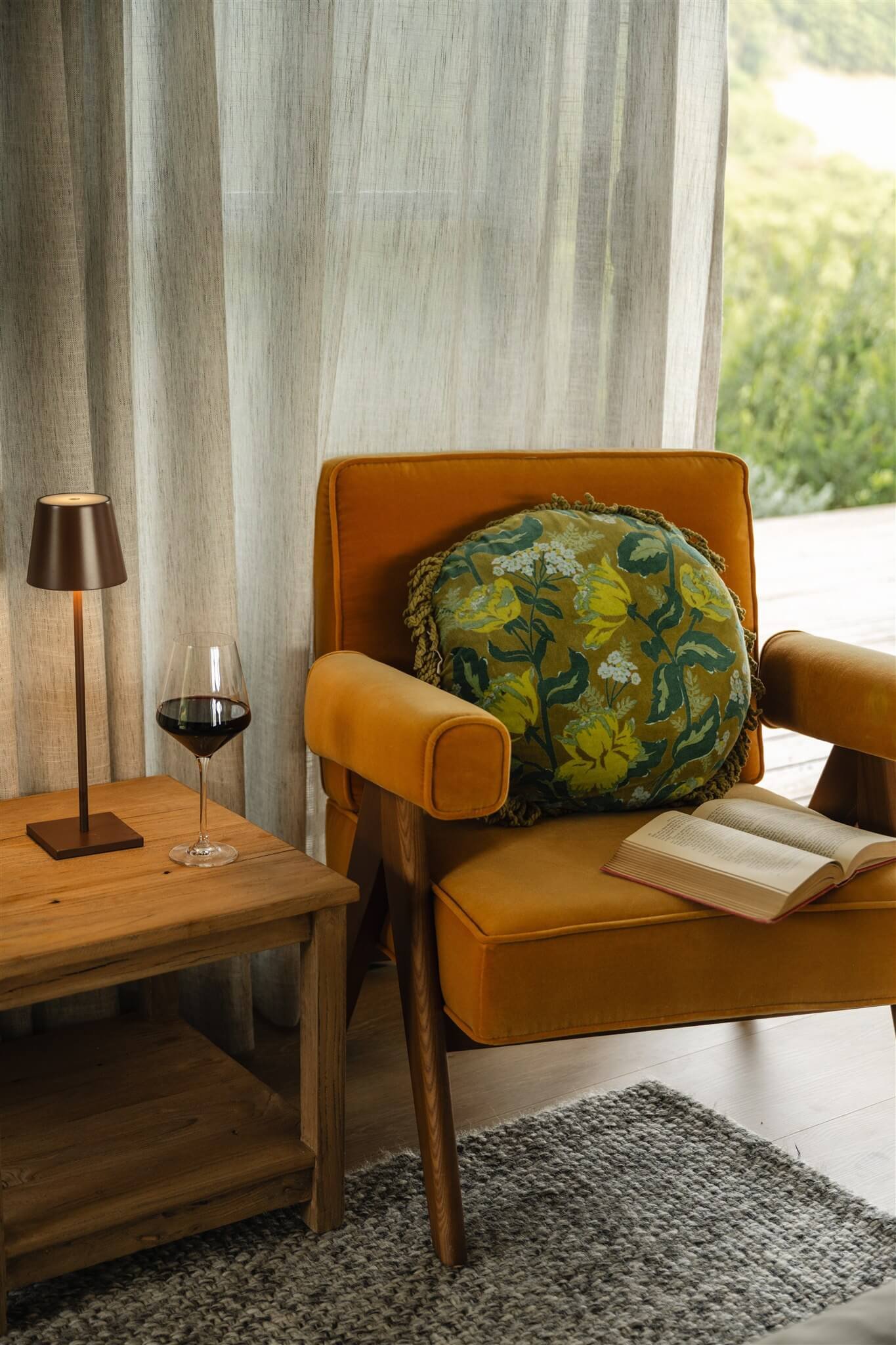 A cozy interior corner with a mustard-colored armchair, a round patterned pillow, a wooden side table with a black lamp, a wine glass filled with red wine, and an open book. A sheer curtain and greenery are visible outside a window.