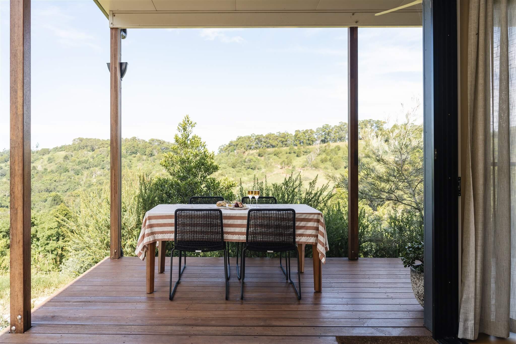 The wooden deck at Marys at Sunshine Coast Hinterland with a table covered by a beige and white striped tablecloth, set with two glasses of white wine, overlooking lush green hills and trees under a blue sky.