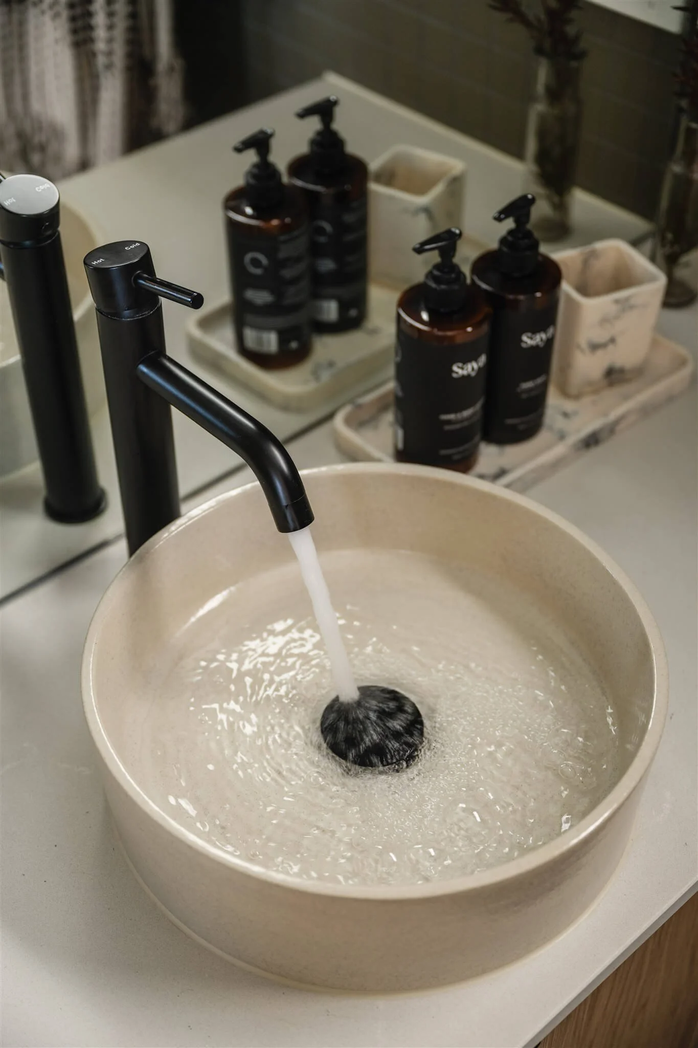 A beige sink with a black faucet running water into it, positioned on a light-colored countertop. In the background, there are several brown soap dispensers and small white containers on a tray, with a mirror and some vases with flowers reflected beh