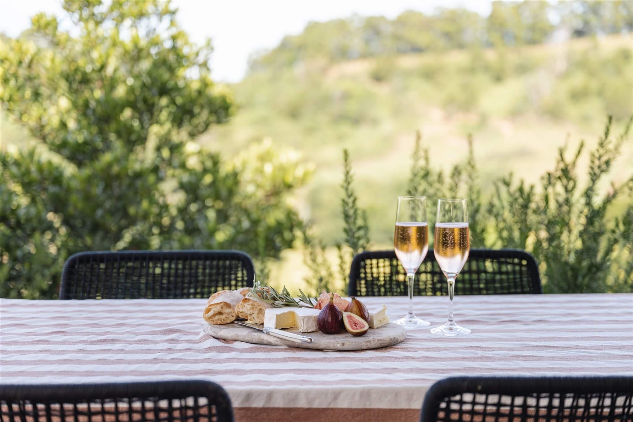 Outdoor table at Marys at Sunshine Coast Hinterland with cheese, figs, bread, and two glasses of champagne, set against a green, hilly landscape.