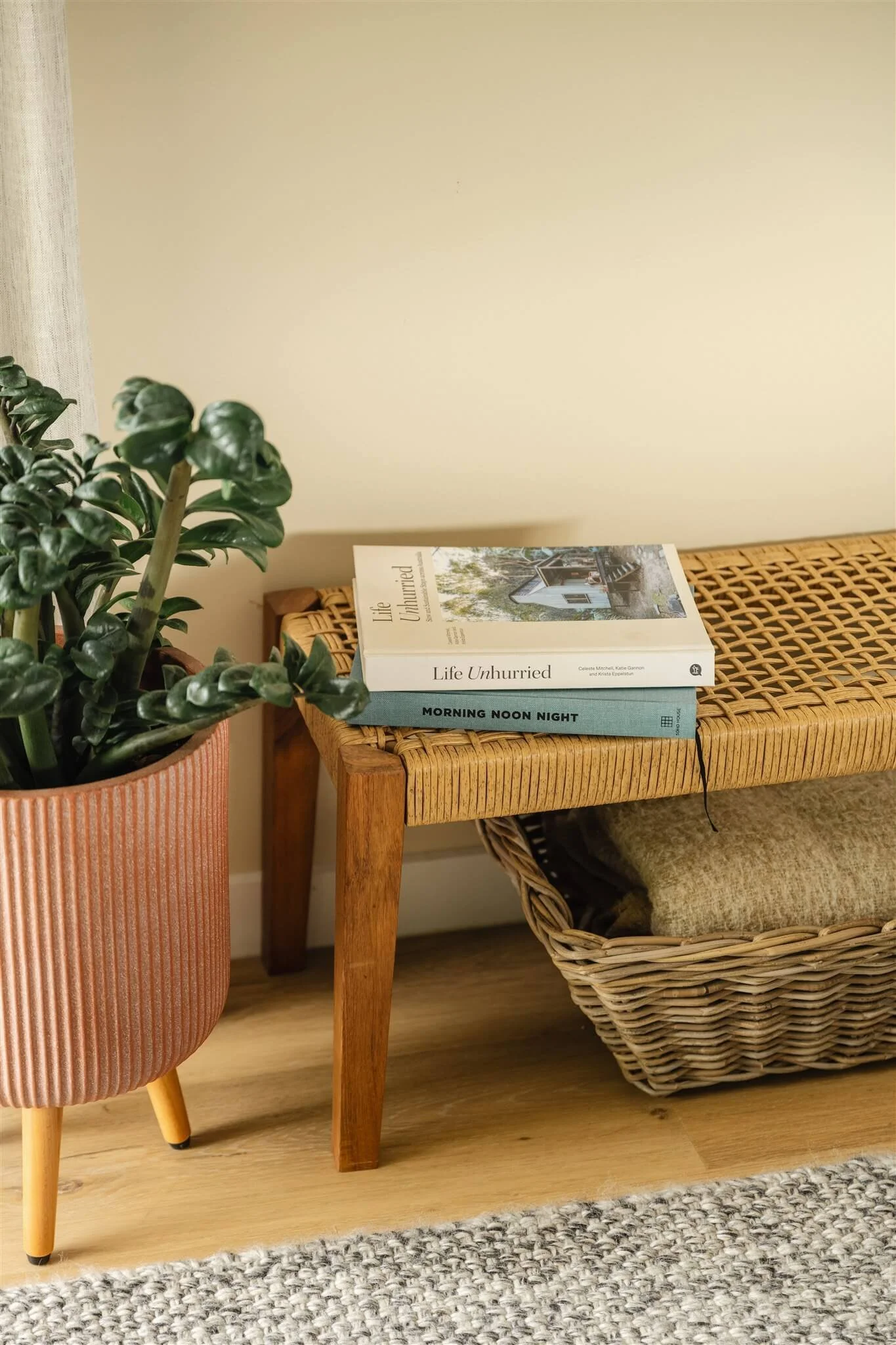 A potted green plant on a pink ribbed pot, a wicker bench with two books on top, one titled 'Life Unhurrried' and the other 'Morning Noon Night,' and a wicker basket with folded fabric underneath, on a wooden floor with a beige wall background.