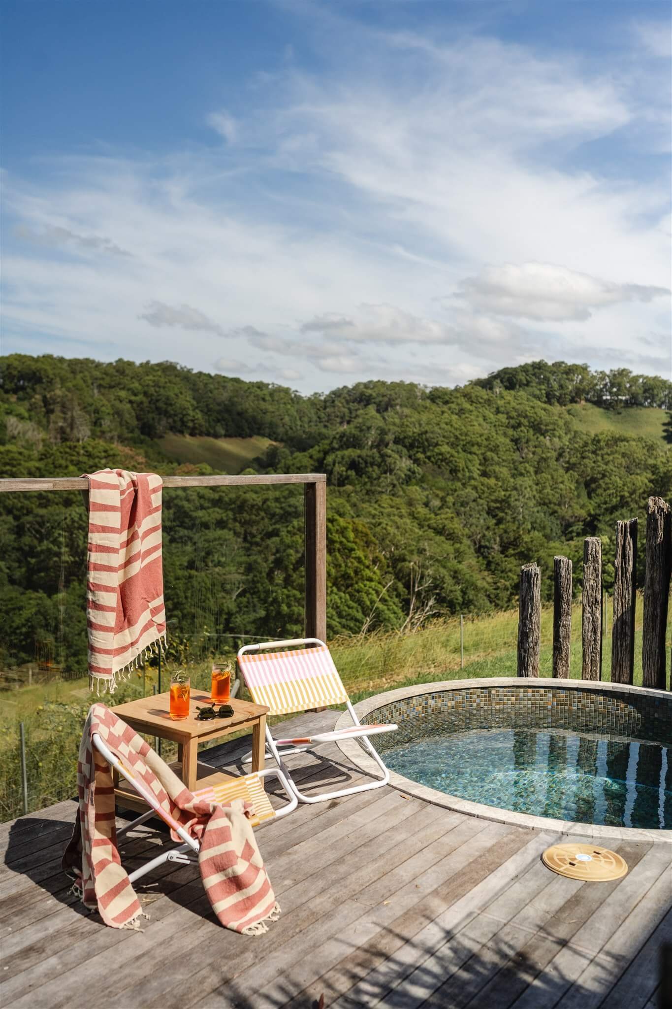 Marys at Sunshine Coast Hinterland's wooden deck with two striped beach chairs, a small wooden table with two glasses of iced tea, sunglasses, and a towel on a railing, overlooking green hills and a blue sky with clouds.