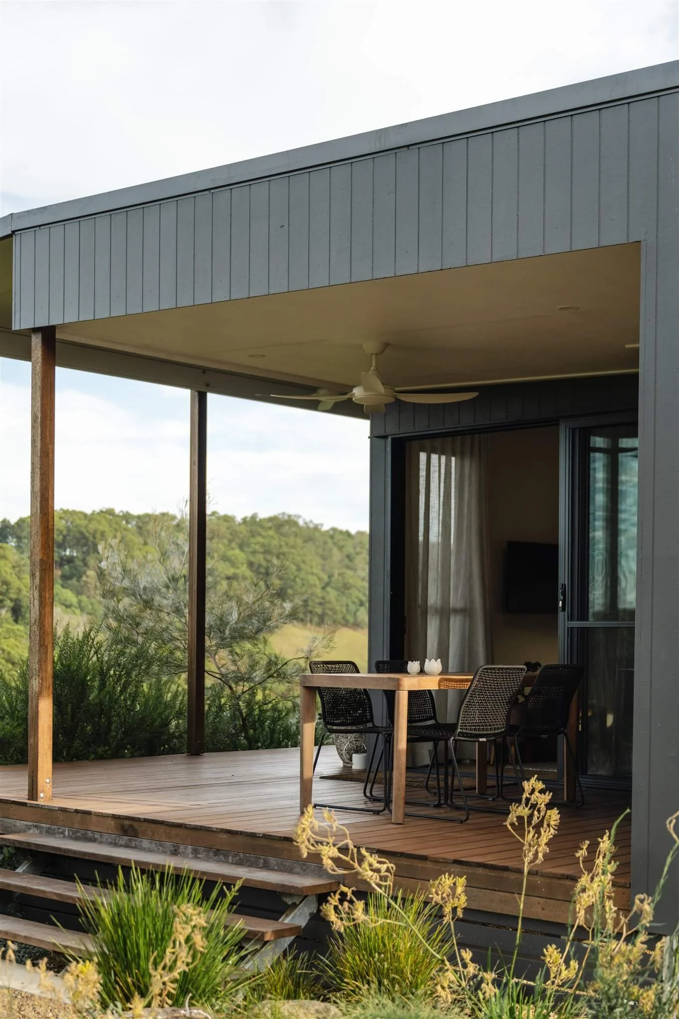 Marys at Sunshine Coast Hinterland. A house with a wooden deck, outdoors, with a round table and four black chairs, greenery, and a ceiling fan underneath a covered patio.