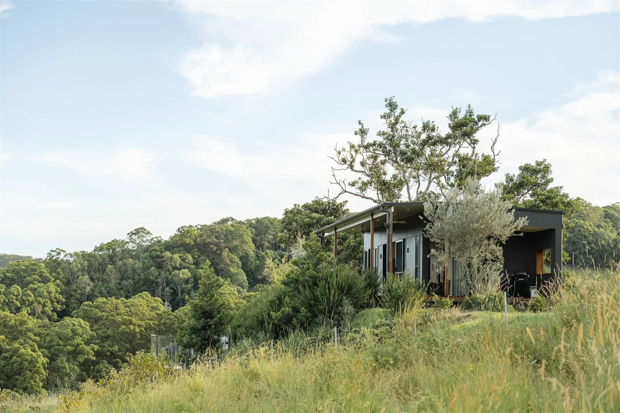 Marys at Sunshine Coast Hinterland is a modern house on a grassy hillside surrounded by trees, with a background of lush forested hills and a partly cloudy sky.